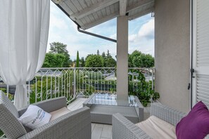 A balcony with wicker furniture, a glass table, and a view of greenery and buildings.