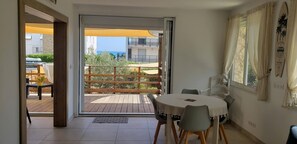A room with a sliding glass door leading to an outdoor deck, a round table with chairs, and a surfboard decoration on the wall. - Duplex Avec Jardin, Piscine, Terrasse, 3 Chambres (Palavas-les-Flots)