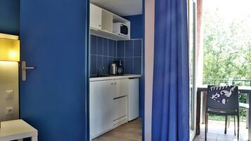 Una cocina compacta con armarios blancos, un microondas y una cafetera. Se ve una silla cerca de una ventana con cortinas azules.