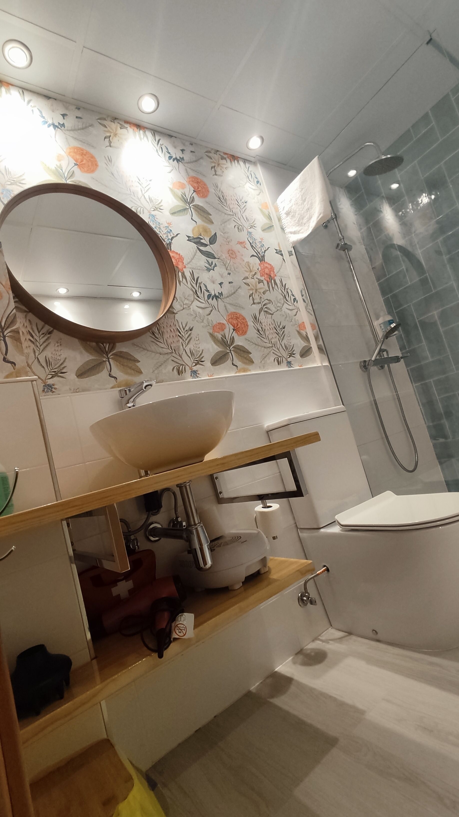 Un baño con papel tapiz floral, un espejo redondo, un lavamanos, una ducha y un inodoro.