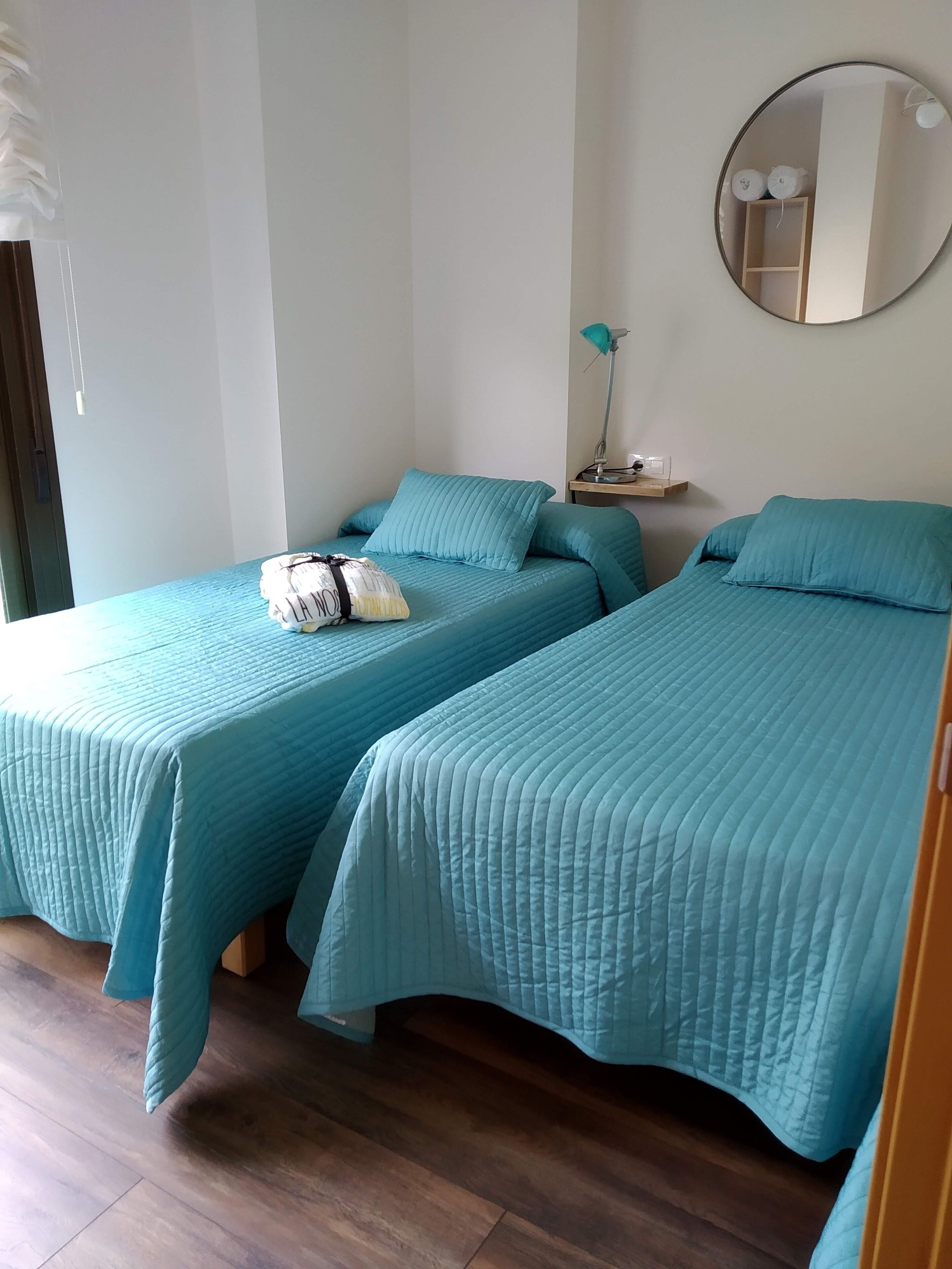Una habitación con dos camas, cada una con ropa de cama y almohadas color turquesa, un espejo redondo en la pared, una mesita con una lámpara y suelo de madera.