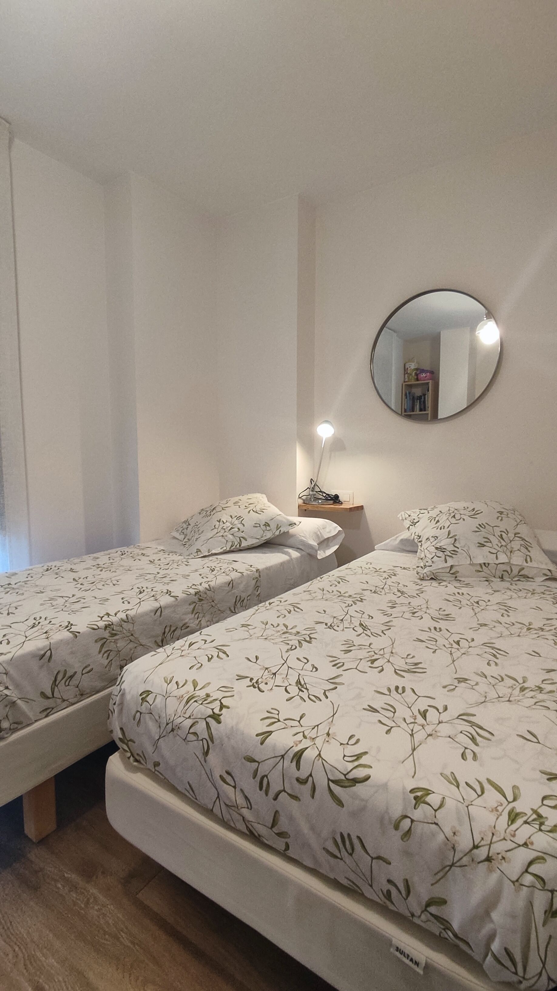 Ett hotellrum med två separata sängar, ett nattduksbord med en lampa, en rund spegel på väggen och ett fönster med gardiner.