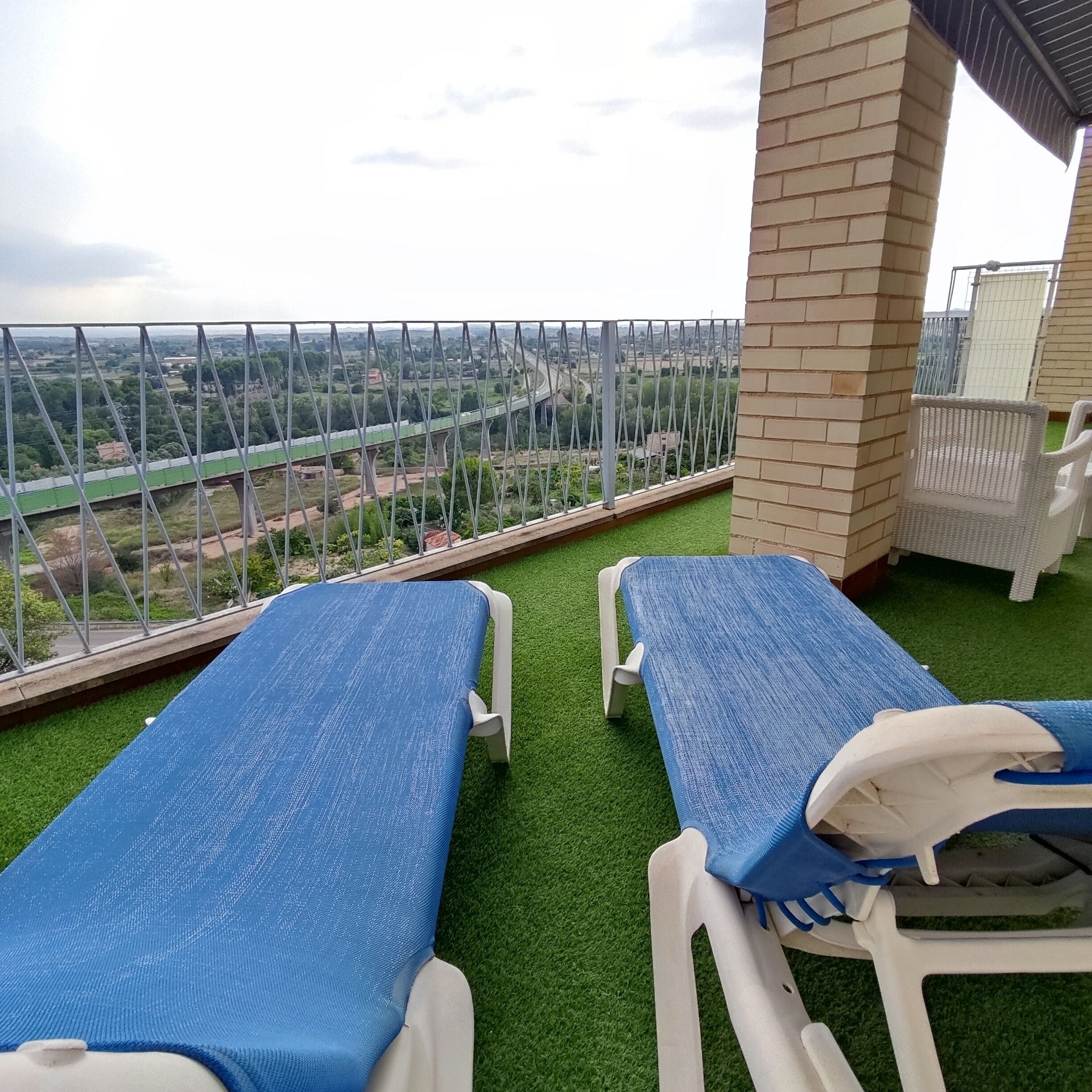 Two sun loungers with blue covers on a balcony with a view of a bridge and greenery.