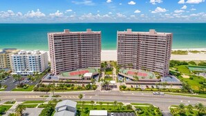 Un hôtel de grande hauteur offrant une vue sur la plage, comprenant de nombreux étages, une cour intérieure avec des courts de tennis et un espace de stationnement.