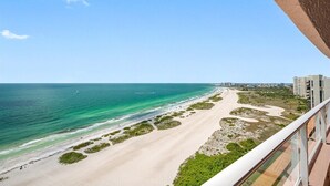 Une vue sur la côte, depuis un balcon donnant sur une plage aux eaux cristallines, avec des bâtiments visibles au loin.