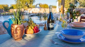 Une table dressée à l’extérieur, avec une bouteille de vin, un saladier de fruits, un vase contenant des plantes vertes et une assiette bleue.