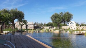 Une terrasse en bois mène à une étendue d’eau calme, avec des structures en pierre et des arbres en arrière-plan.