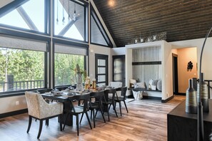 Uma sala de jantar com uma mesa grande, cadeiras e vista para árvores através de amplas janelas.