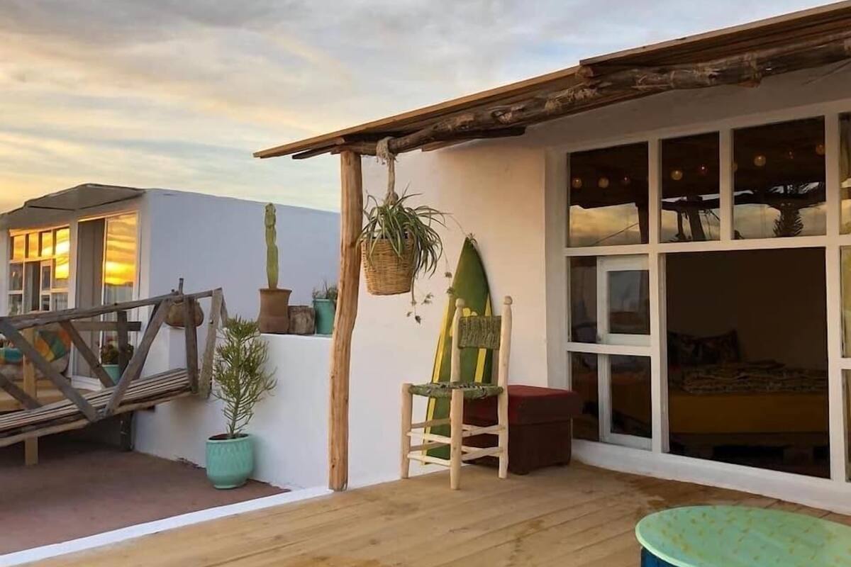 A wooden deck with a table and chairs, a hanging plant, and a large window with a view.