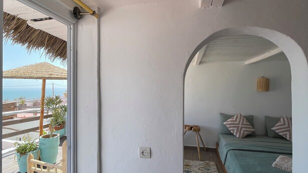 A room with a bed, a wooden chair, a small table, and a view of the sea through a glass door. - Surf house Maroc (Essaouira)