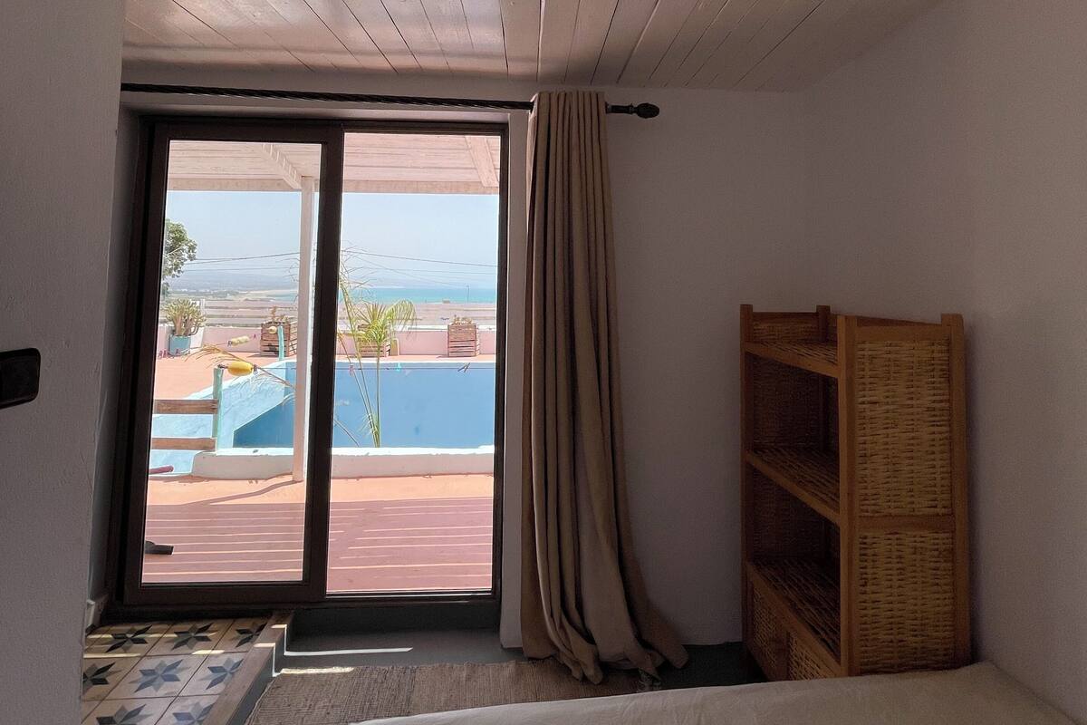 A bedroom with a bed, a wooden shelf, a window with a view of a pool and palm trees, and a sliding door.