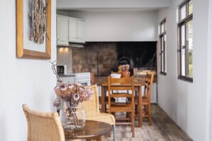 A dining area with a round table, wicker chairs, and a vase with dried flowers. A kitchen with white cabinets and a black backsplash is visible in the background.