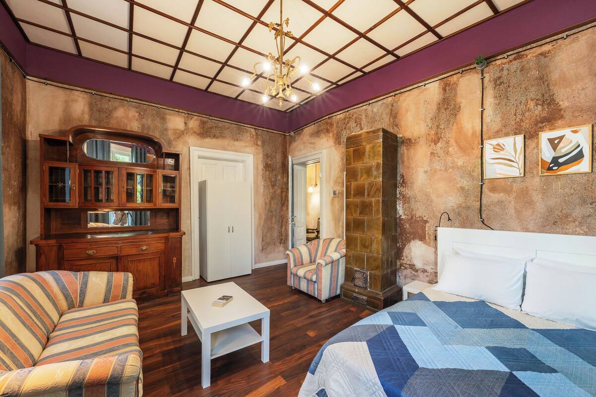 A room with a bed, a striped armchair, a small white table, a wooden cabinet, and a door leading to another room.
