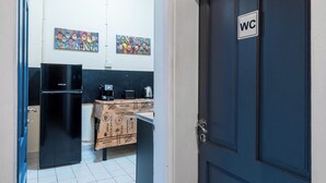 A narrow hallway leading to a kitchen with a refrigerator, a coffee machine, and a table covered with a tablecloth. There are two paintings on the wall and a door labeled "WC". - The Green Bar House (Sofia)