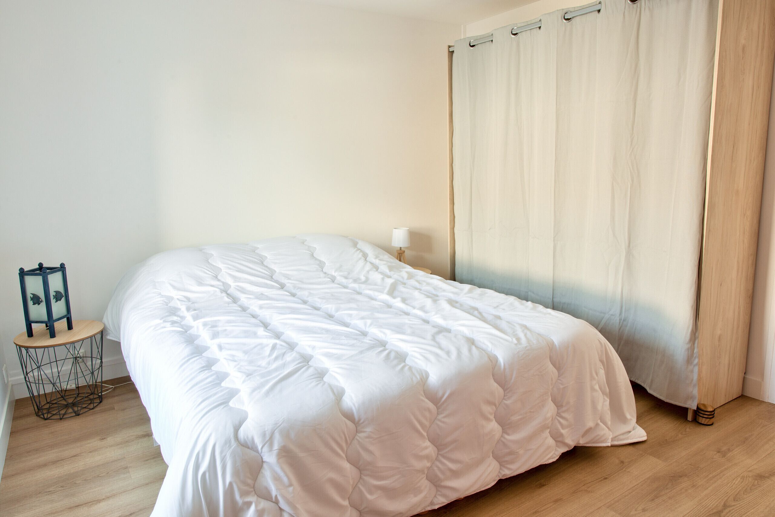 A neatly made bed in a room with a wooden floor, a small round table with a lamp, and a curtain on the window.