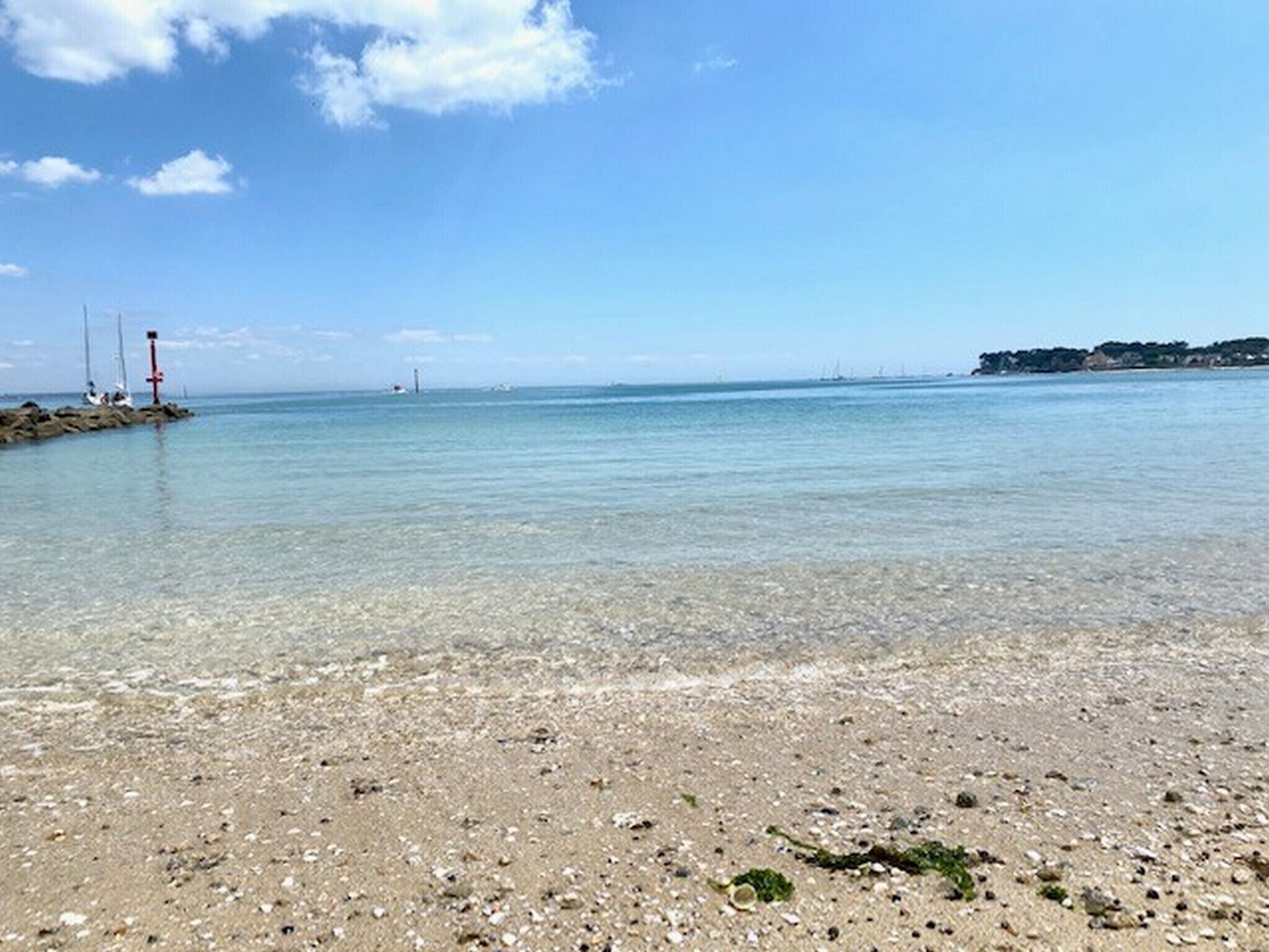 A beach with clear shallow waters, a rocky shoreline, and a distant view of a lighthouse and sailboats.