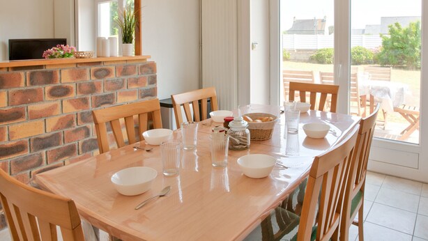A dining area with a brick wall, wooden chairs, a glass table, and a view of the outside. - Large family home with garden- For 10 (Penmarch)