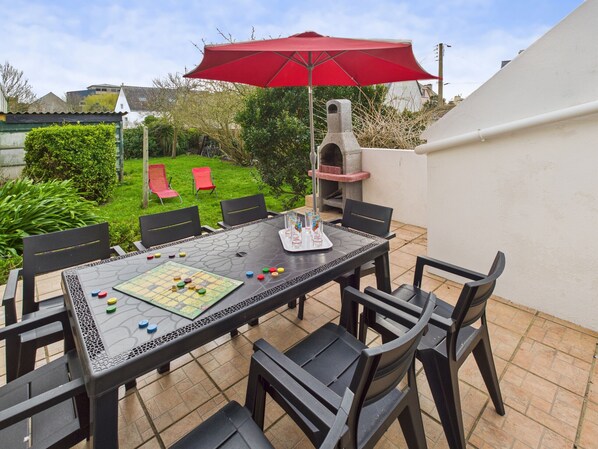 Um pátio externo com uma mesa preparada para um jogo de tabuleiro, rodeado por cadeiras, sob um guarda-sol vermelho.