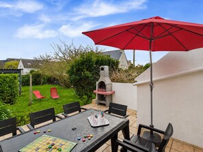 Um pátio externo com mesa preparada para um jogo, um guarda-sol vermelho e uma churrasqueira.