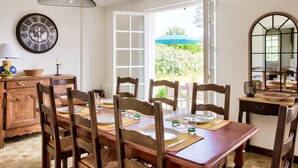 Comedor con mesa de madera dispuesta para seis personas, rodeada de sillas, un reloj y una ventana con vistas a zonas verdes.