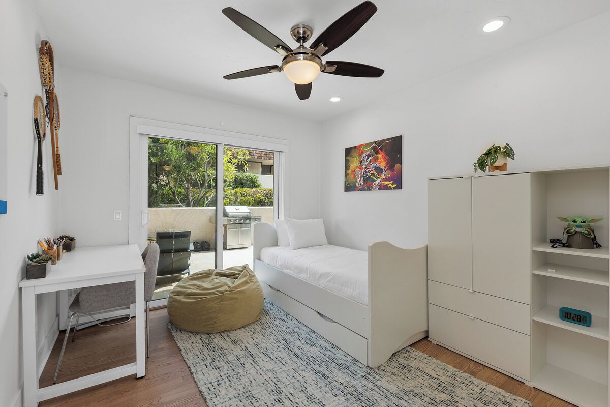 Une chambre à coucher de taille modeste, comprenant un lit, un bureau et une armoire, et dotée d’un ventilateur de plafond et d’une vue sur la verdure environnante.