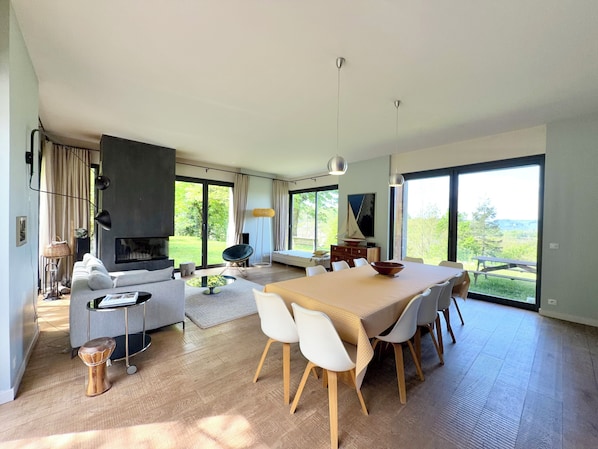 A modern living room with a dining area, large windows, and a fireplace.