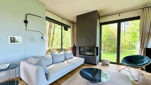 Een moderne woonkamer met een bank, een glazen salontafel, een open haard en grote ramen met uitzicht op groen.