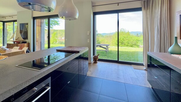 Een moderne keuken met een groot eiland, een eethoek met een bank en uitzicht op een gazon door grote ramen.