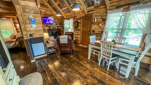 L’intérieur chaleureux d’un chalet avec un plancher en bois, une table à manger avec des chaises, une cheminée et un téléviseur fixé au mur.