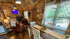 L’intérieur chaleureux d’un chalet avec une table à manger en bois, des chaises blanches, une cheminée et un téléviseur fixé au mur.