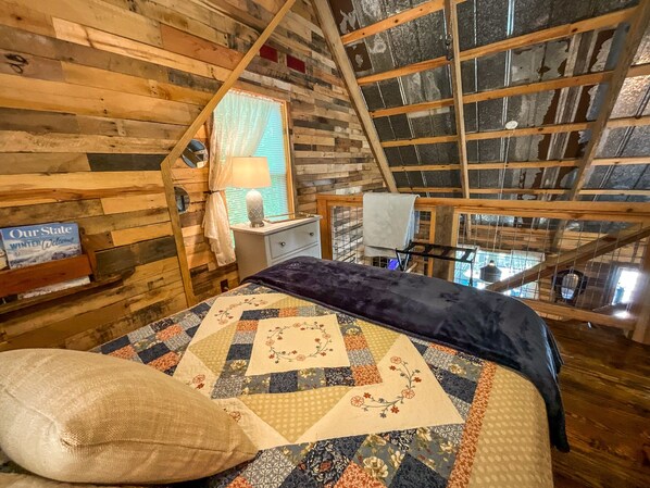 Een knus kamer in een blokhut, met houten inrichting, een bed met een bloemenquilt, een nachtkastje en een raam met gordijnen.