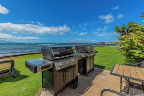 An outdoor grill area with a view of the ocean, a grassy lawn, and a wooden table.