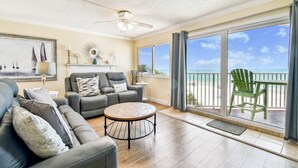 A spacious living room with a large window offering a view of the beach, a ceiling fan, and a round coffee table.