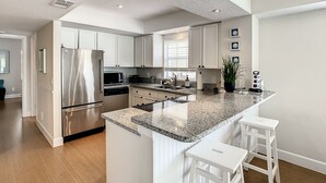 Una cocina moderna con armarios blancos, un refrigerador de acero inoxidable, una encimera de granito y una zona de bar con taburetes.