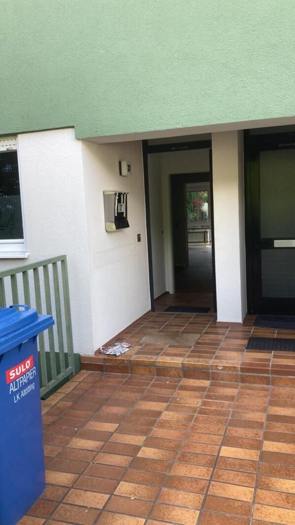 A covered outdoor area with a trash bin, a bench, and a door leading to an interior space. - ZV-Scholtz Burghausen Regerstr.37 (Burghausen)