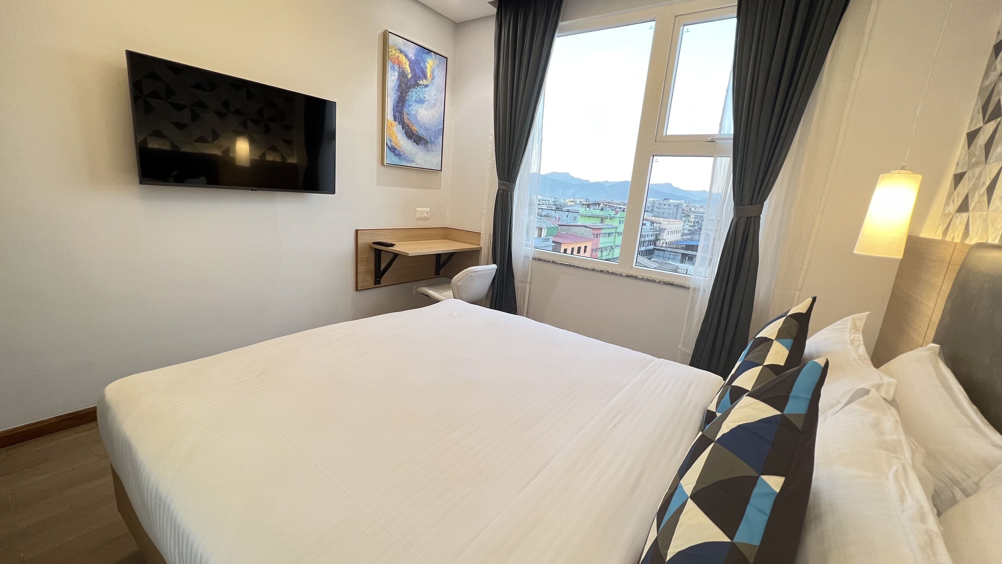Habitación de hotel con una cama grande, un escritorio, una silla, un televisor y ventana con vista a la ciudad.