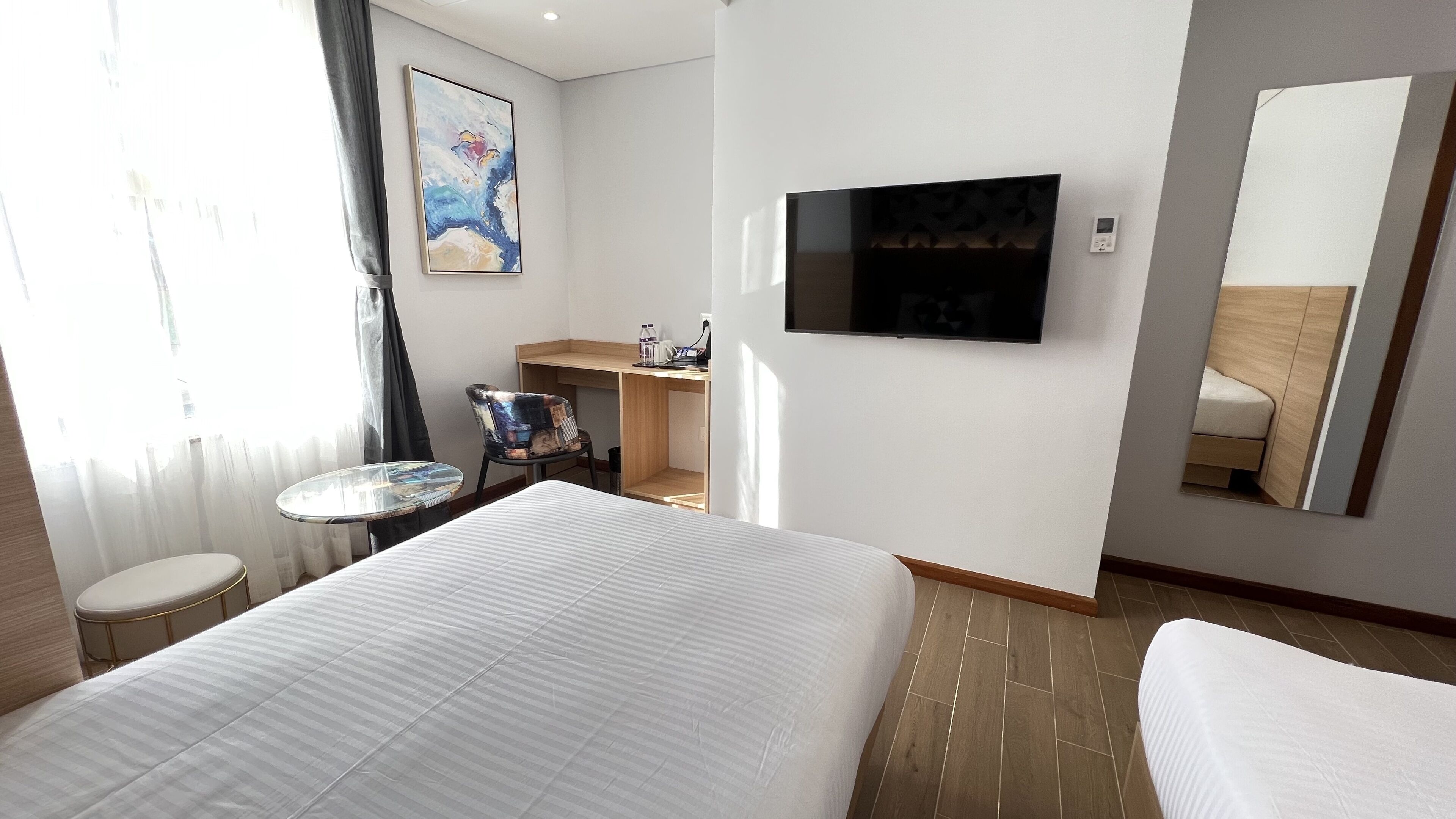 Hotellihuone, jossa on suuri sänky, pieni pyöreä pöytä, työpöytä tuolilla, seinälle asennettu televisio ja suuri peili.