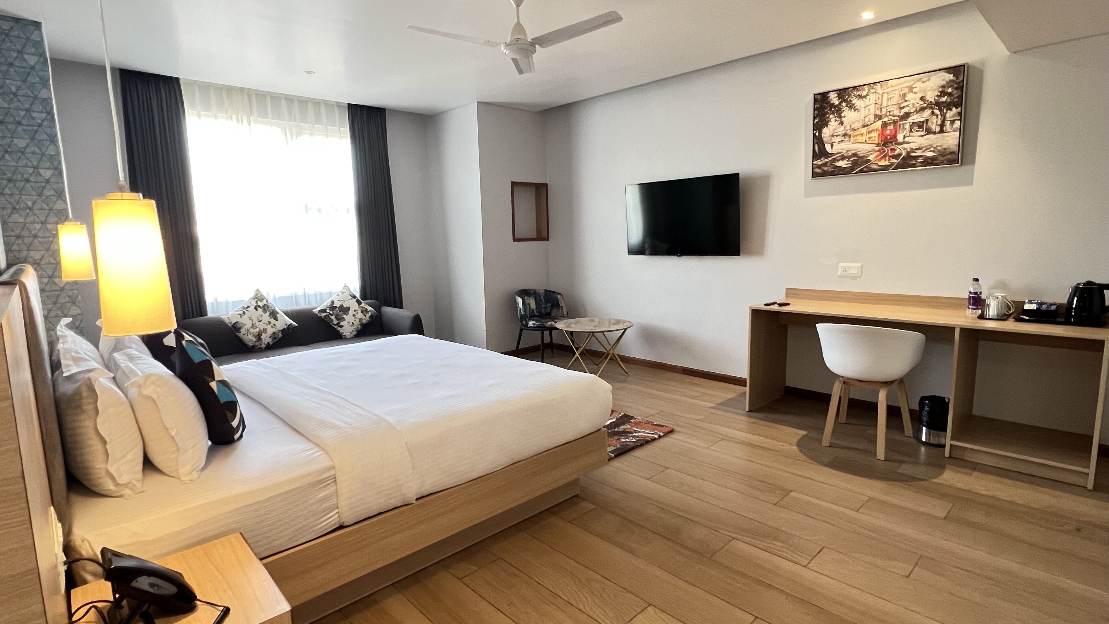 A modern hotel room with a bed, desk, chair, TV, and artwork on the wall.