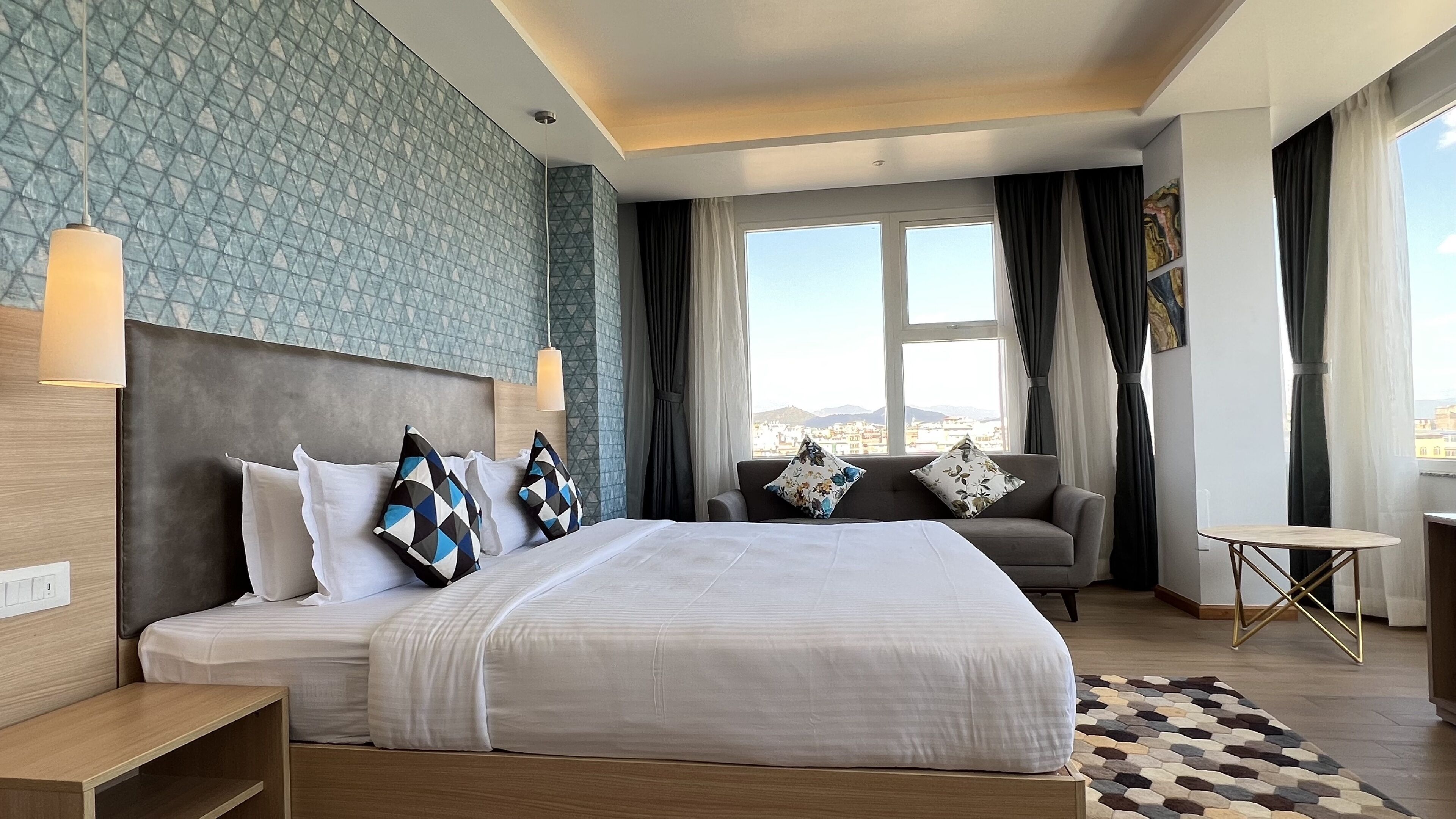 A modern hotel room with a large bed, a sofa, a small table, and a view of the city.