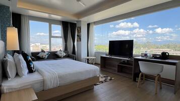 A modern hotel room with a large bed, a desk with a flat-screen TV, and a view of the cityscape through large windows.