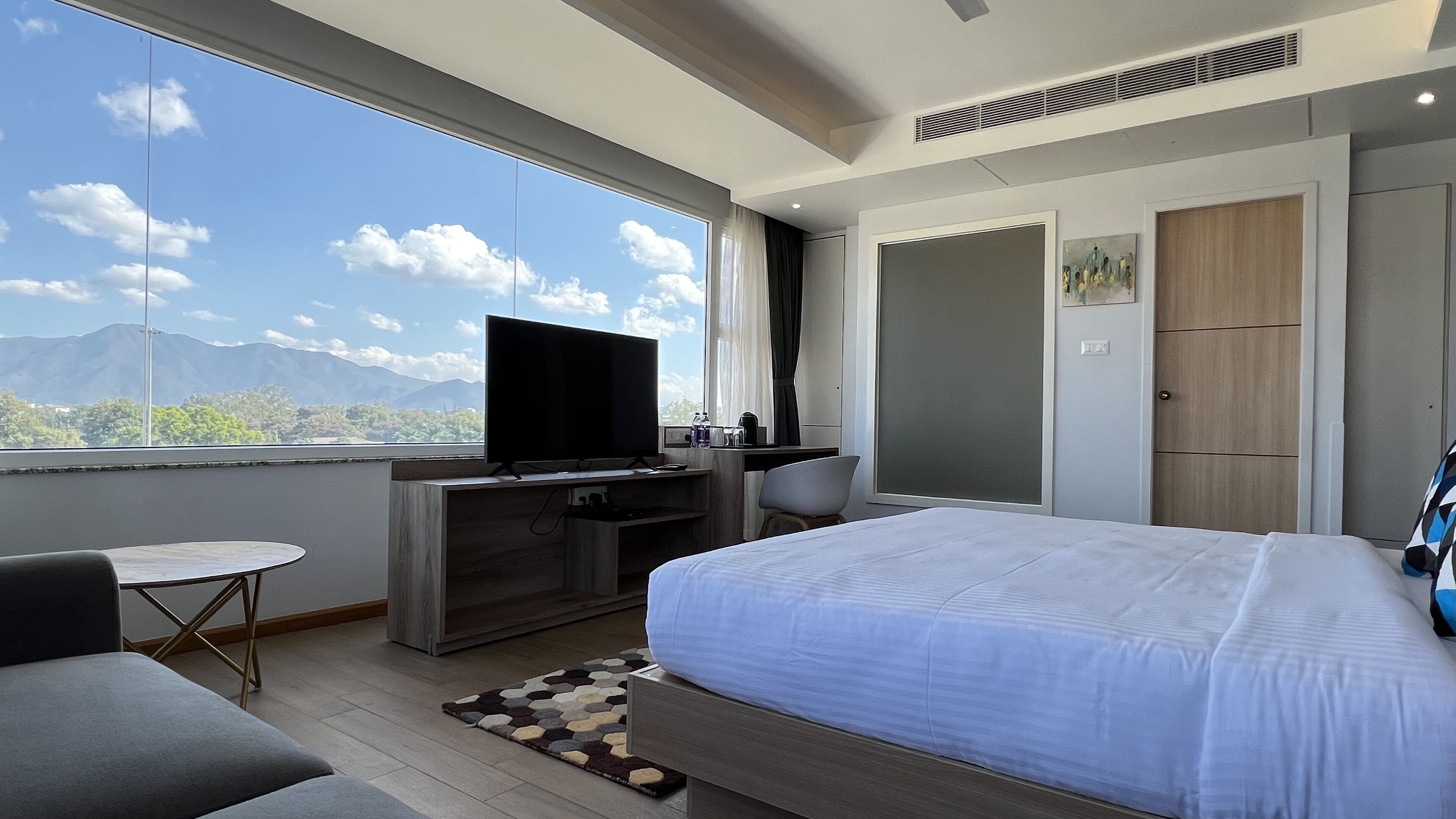 A modern hotel room with a large bed, a desk with a TV, a small table, and a view of mountains through the window.