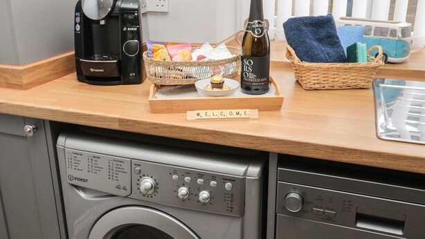 Una encimera de cocina con una cafetera, una botella de vino, una cesta de aperitivos y una bandeja con un juego de Scrabble.
