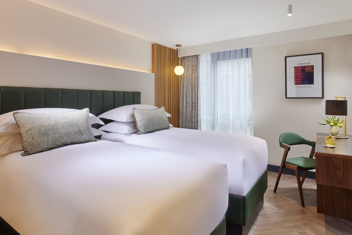Ein Hotelzimmer mit zwei Betten, einem Schreibtisch, einem Sessel, einer Lampe und einem gerahmten Bild an der Wand.
