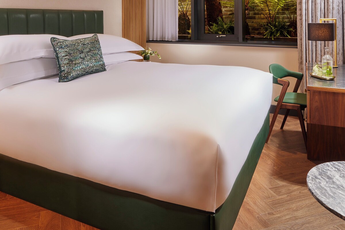 A modern hotel room with a large bed, a desk, and a view of greenery through the window.
