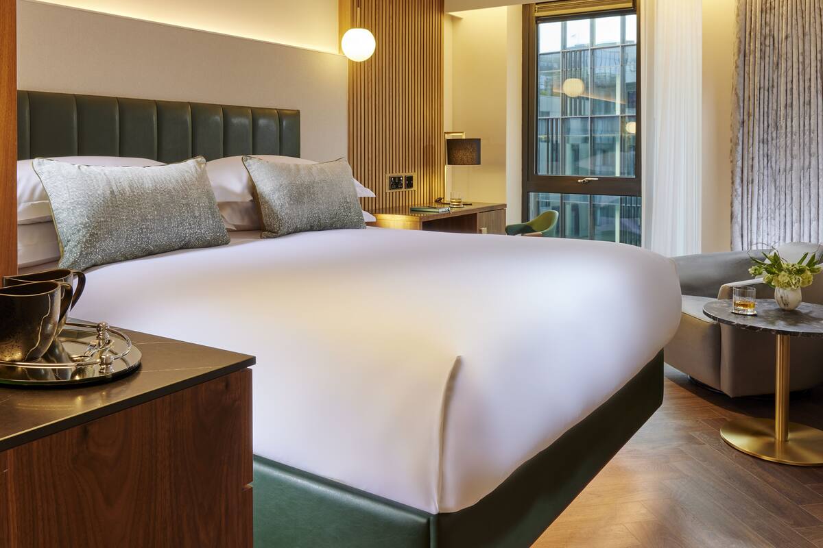 Ein modernes Hotelzimmer mit einem großen Bett, einem Nachttisch mit Lampe, einem kleinen Schreibtisch, einem Sessel und Blick auf die Stadt durch große Fenster.