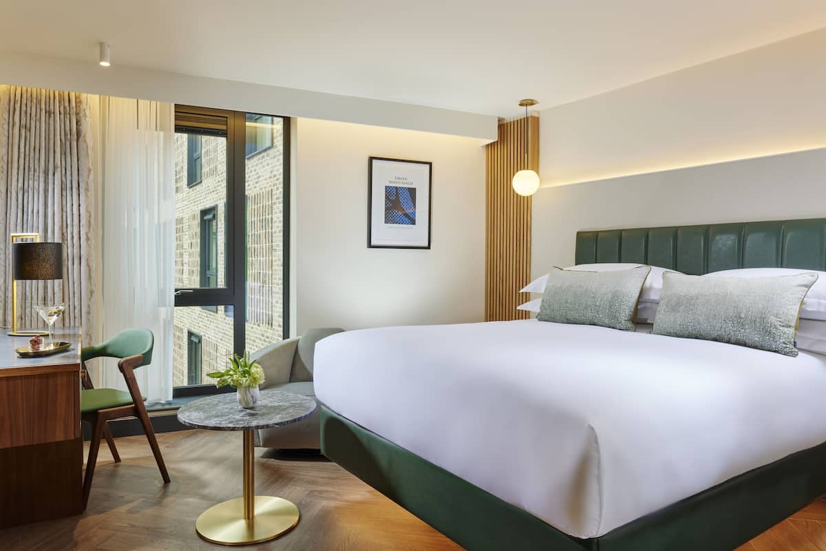 Ein Hotelzimmer mit einem großen Bett, einem Schreibtisch mit Stuhl, einem kleinen runden Tisch und einem Fenster mit Blick auf ein Backsteingebäude.