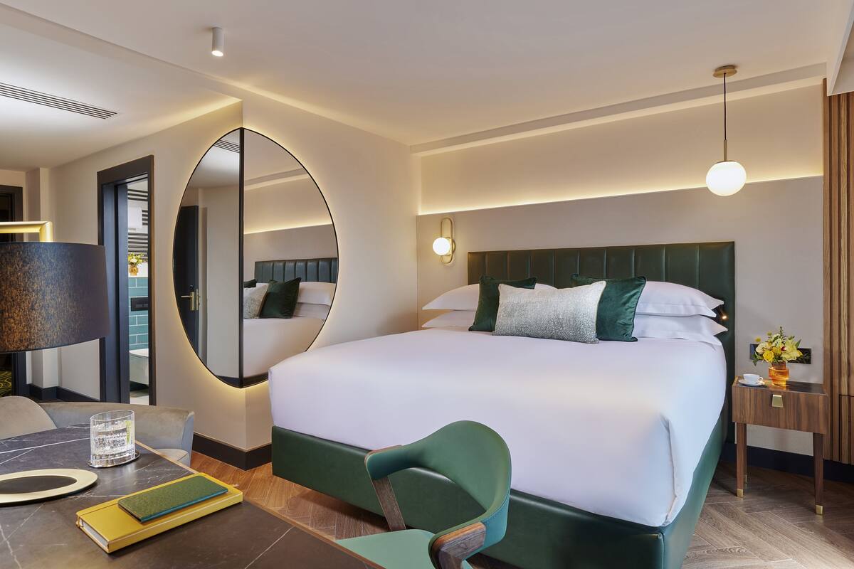 Ein modernes Hotelzimmer mit einem großen Bett, einem runden Spiegel, einem Schreibtisch mit Lampe und einem kleinen Tisch mit Blumenvase.