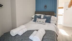 A neatly made bed with white linens and rolled towels, a blue accent wall, and a modern pendant light.
