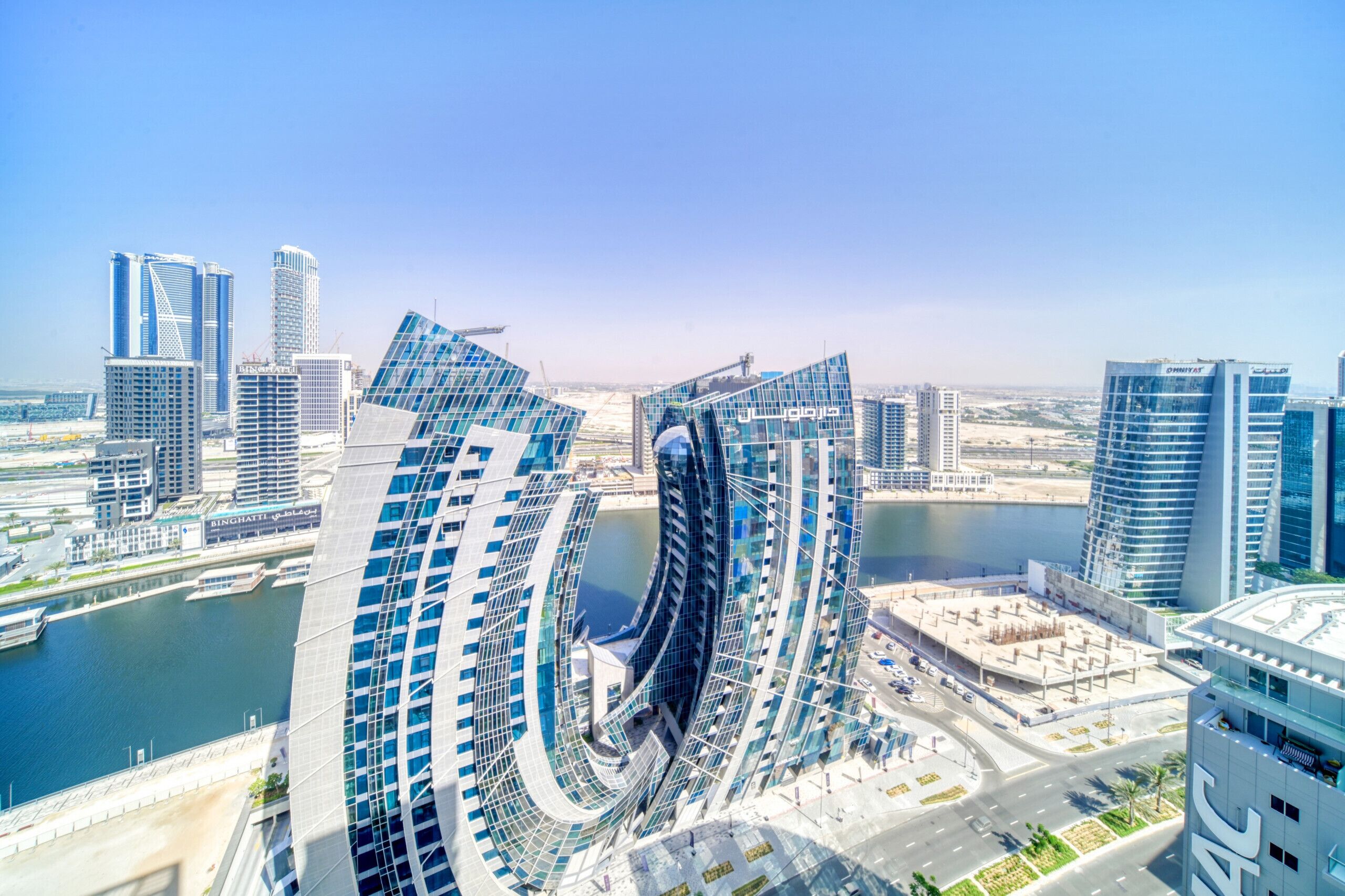 Un paysage urbain moderne, avec un bâtiment aux lignes courbes originales, entouré d’autres immeubles de grande hauteur et bordé par une étendue d’eau.