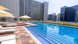 Una piscina con lettini e ombrelloni, circondata da edifici moderni.